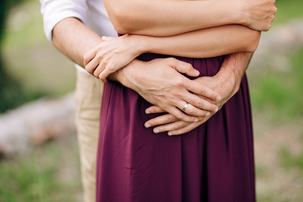 Τα χέρια του γαμπρού σε ένα λευκό πουκάμισο αγκαλιά από πίσω νύφη σε ένα μπορντό φόρεμα. Κοντινό πλάνο - Φωτογραφία, εικόνα