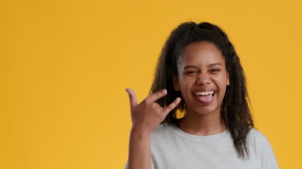 Czarny nastolatek dziewczyna Gesturing skała znak Pokazuje język, żółte tło - Materiał filmowy, wideo