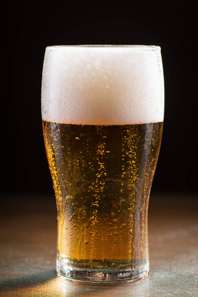 A vertical shot of a glass of cold refreshing beer with foam on a wooden table - Фото, изображение