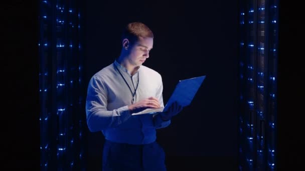 Ειδικός πληροφορικής εργάζεται με ένα φορητό υπολογιστή στην αίθουσα του διακομιστή κοιτάζοντας τα ντουλάπια με διακομιστές - Πλάνα, βίντεο