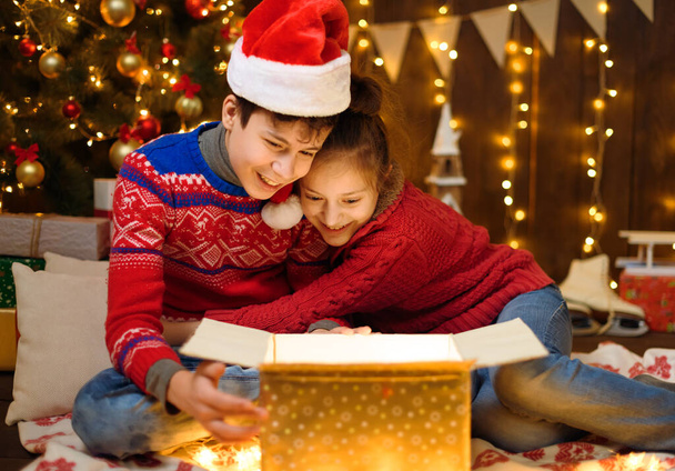 Портрет мальчика и девочки в новогоднем оформлении. Они открывают коробку и веселятся. Рождественские огни, подарки и елка, украшенные игрушками. - Фото, изображение