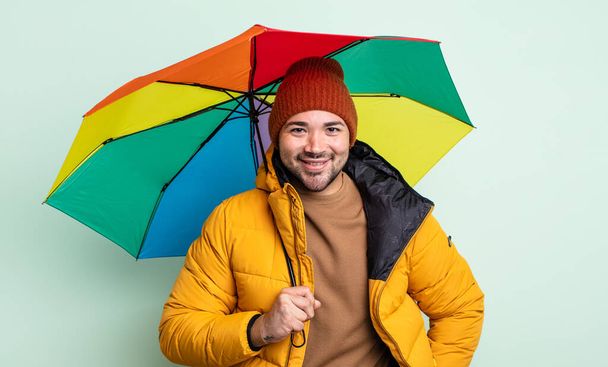 young handsome man smiling happily with a hand on hip and confident. rain and umbrella concept - Фото, изображение