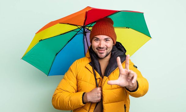 young handsome man smiling and looking friendly, showing number three. rain and umbrella concept - Fotografie, Obrázek