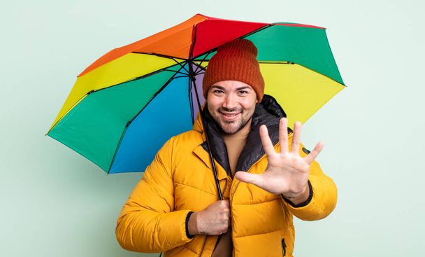 young handsome man smiling and looking friendly, showing number five. rain and umbrella concept - Fotoğraf, Görsel