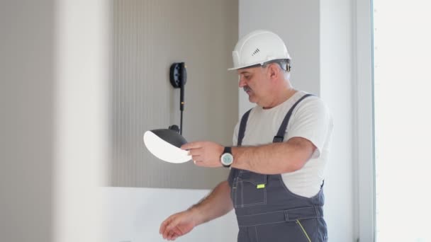 Un electricista de edad avanzada comprueba el funcionamiento de los apliques de pared de la lámpara en la casa después de la instalación y reparación - Imágenes, Vídeo