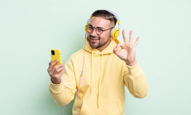 young handsome man feeling happy, showing approval with okay gesture. headphones and smartphone concept - Фото, изображение