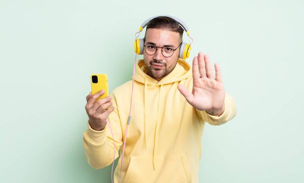 young handsome man looking serious showing open palm making stop gesture. headphones and smartphone concept - Foto, Imagem
