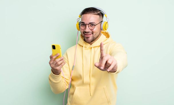 young handsome man smiling proudly and confidently making number one. headphones and smartphone concept - Fotografie, Obrázek