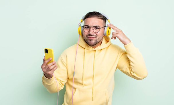 young handsome man feeling confused and puzzled, showing you are insane. headphones and smartphone concept - Zdjęcie, obraz