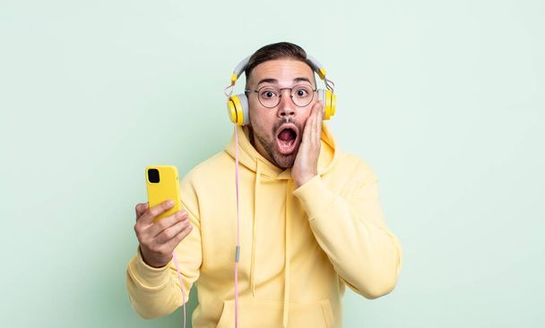 young handsome man feeling shocked and scared. headphones and smartphone concept - Foto, Imagen