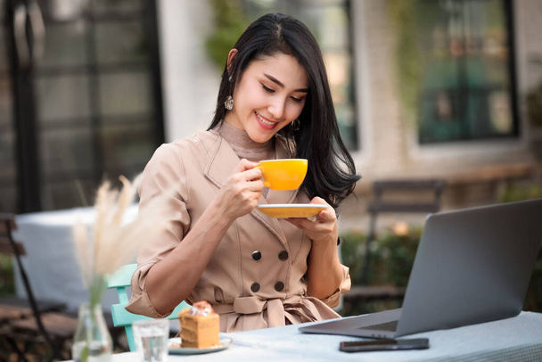 Happy young woman is drinking hot beverage and cake at workplace. She is looking at cup with joy and smiling - Photo, Image