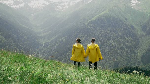 Po svahu sestupují mladý muž a žena ve žlutých pláštích, drží se za ruce a užívají si scenérie hornaté oblasti. Cestování a cestovní ruch - Fotografie, Obrázek