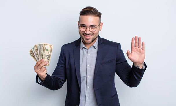 young handsome man smiling happily, waving hand, welcoming and greeting you. dollar banknotes concept - Fotoğraf, Görsel