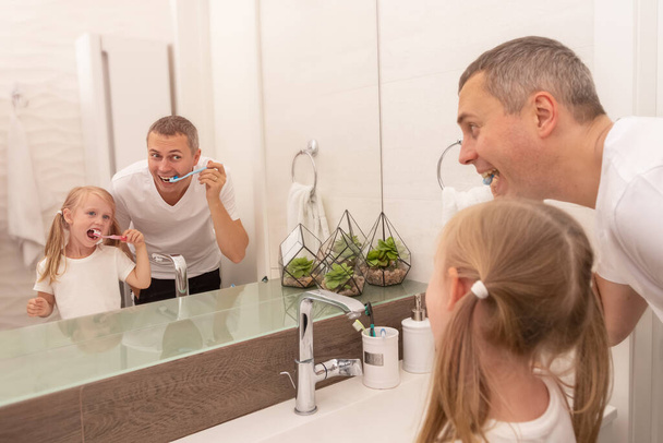 Dad teaches daughter to brush teeth with toothbrush and toothpaste in bathroom - Фото, зображення
