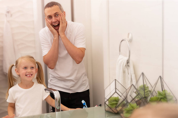 Daughter shows father fallen milk tooth reflected in mirror in bright bathroom - Fotoğraf, Görsel