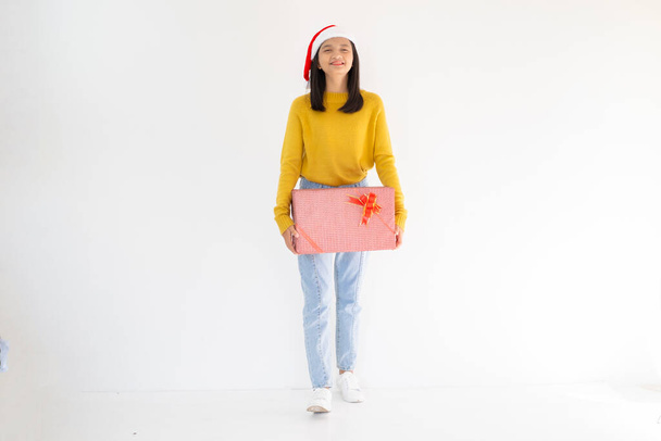 Счастливая молодая девушка держит подарочную коробку, азиатская девушка носит желтый свитер на белом фоне. - Фото, изображение