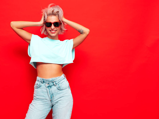 Young beautiful smiling female in trendy summer clothes. Sexy carefree woman posing near red wall in studio. Positive blond model having fun and going crazy. Cheerful and happy. In sunglasses - Fotoğraf, Görsel