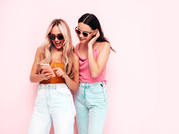 Dwie młode, piękne, uśmiechnięte brunetki hipsterki w modnych letnich ciuchach. Seksowne beztroskie kobiety pozujące przy różowej ścianie. Pozytywne modelki dobrze się bawią. Wesoły i szczęśliwy. Trzymanie smartfona. Korzystanie z aplikacji - Zdjęcie, obraz