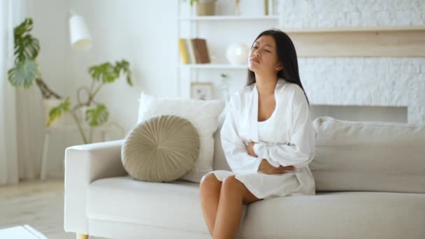 Douleur féminine. Jeune femme asiatique souffrant de douleurs menstruelles, tenant l'estomac douloureux sur le canapé à la maison, espace libre - Séquence, vidéo