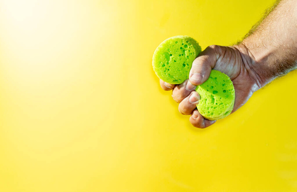 Αρσενικό χέρι κρατά και σφίγγει ένα πράσινο σφουγγάρι, απομονωμένο σε κίτρινο φόντο.  - Φωτογραφία, εικόνα