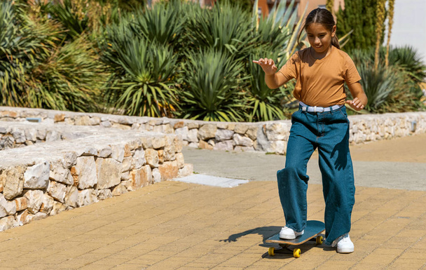 Девочка в бежевой футболке учится кататься на скейтборде на открытом воздухе. Милый ребенок занимается спортом и веселится в парке. Активные увлечения, концепция здорового образа жизни. Макет футболки - Фото, изображение
