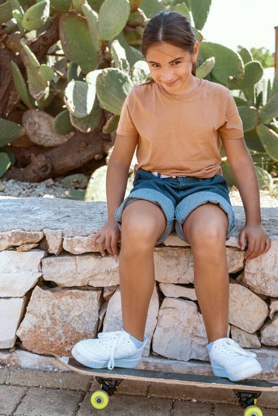 Écolière en t-shirt assise avec une planche à roulettes à l'extérieur. Enfant mignon faisant du sport et s'amusant dans le parc. Loisirs actifs, concept de mode de vie sain. Modèle de T-shirt - Photo, image