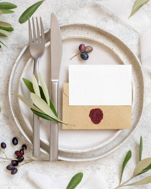 Свадебный стол с карточкой и конвертом поверх фарфоровых тарелок, с оливковыми ветвями вид сверху. Элегантный современный шаблон с горизонтальной пустой бумажной карточкой. Средиземноморский макет - Фото, изображение
