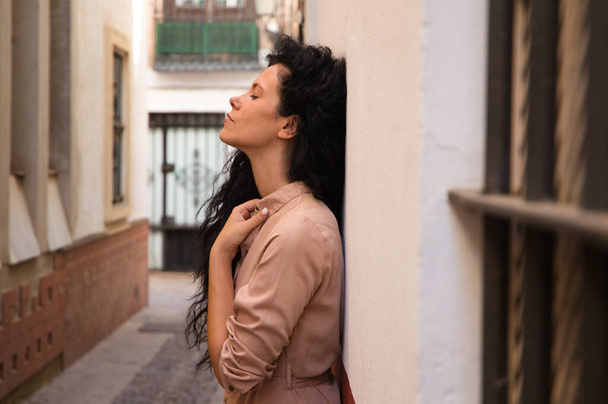 beautiful woman with dark curly hair leaning against the wall of a typical european house in a mediterranean style city. The woman is serious and sad. Concept expressions. - 写真・画像
