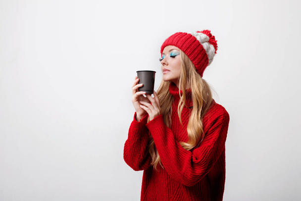 a girl with bright makeup in a winter sweater and a hat with a craft cup of coffee on a gray background - Foto, imagen