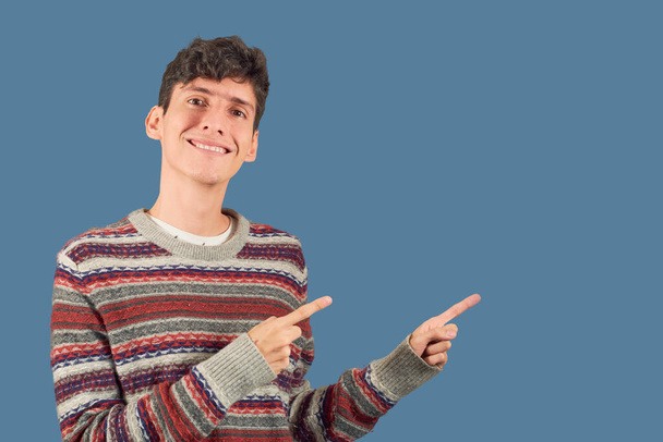 Młody mężczyzna w swetrze wskazuje palcami na boku na płaskim tle z przestrzenią do kopiowania. - Zdjęcie, obraz