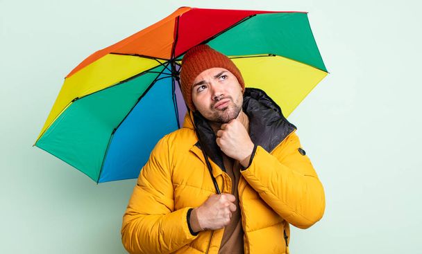νεαρός όμορφος άντρας σκέφτεται, αισθάνεται αμφίβολος και μπερδεμένος. έννοια βροχή και ομπρέλα - Φωτογραφία, εικόνα