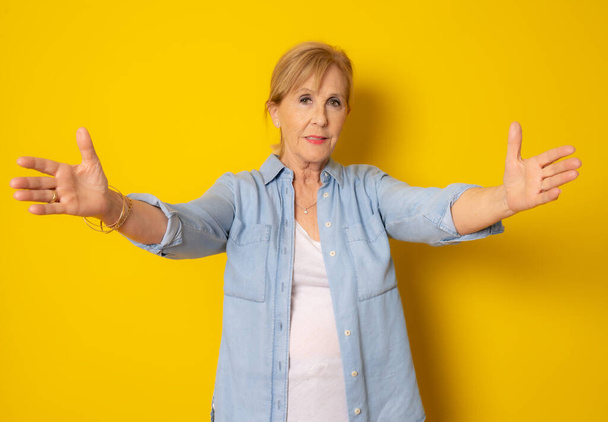 Mature woman in casual clothing giving a hug to camera isolated over yellow background. - Foto, Imagem