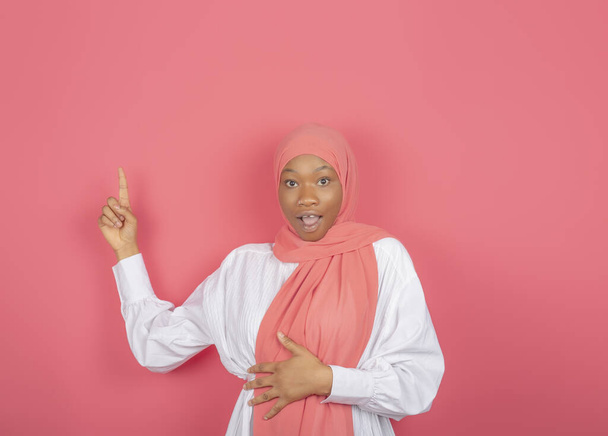 Συναρπαστική νεαρή μουσουλμάνα γυναίκα του χρώματος δείχνει δείκτη δάχτυλο σε ελεύθερο χώρο τυλιγμένο με ροζ πέπλο απομονώνονται σε ροζ φόντο. - Φωτογραφία, εικόνα
