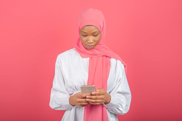 ピンクの背景に隔離されたスマートフォンを使ってアフリカ系アメリカ人の若いイスラム教徒の女性。若いです男でピンクヒジャーブタイピングメッセージで携帯電話. - 写真・画像