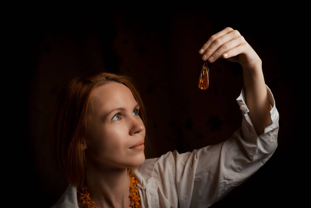 An attractive woman holds a piece of jewelry in her hand - amber pendant on a black background. - Φωτογραφία, εικόνα