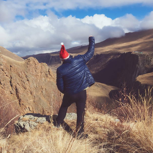 Ein Mann mit Nikolausmütze. Erhob eine Hand, freute sich in der Natur. Vor dem Hintergrund einer großen, schönen Schlucht und Wolken, die auf den Gipfeln der Berge liegen. Weihnachtsposter oder Postkarte - Foto, Bild