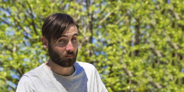 Portrait of a surprised young Caucasian man in close-up. Bearded hipster guy with bangs on the background of green foliage of trees in the park. - Foto, Imagen