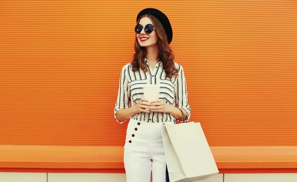 Portrait of beautiful young woman with shopping bags wearing a white striped shirt, black round hat on orange background - Fotografia, imagem