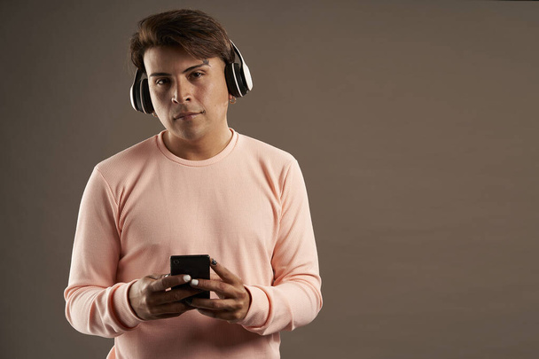 hombre latino con auriculares y teléfono celular en la mano sobre fondo gris y ropa rosa - Foto, imagen