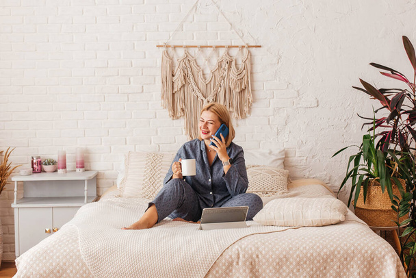 Beautiful young woman in pajamas, working on a laptop in bed, talking on the phone. Online shopping - Foto, Imagem
