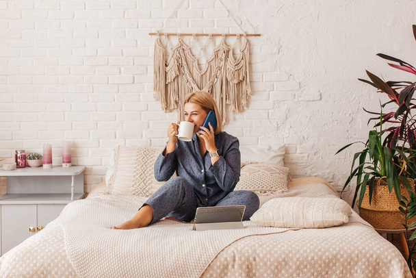 Beautiful young woman in pajamas, working on a laptop in bed, talking on the phone. Online shopping - Фото, изображение