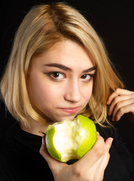 Blondes in black T-shirts on a black background. Eat a green apple. A bone of contention is the core, or the core of the dispute, or a small issue that can lead to a larger dispute. - Foto, Imagen