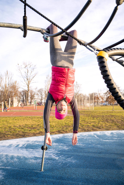Young fit woman in fitness clothes hanging with legs upside down on sports bar - Fotó, kép