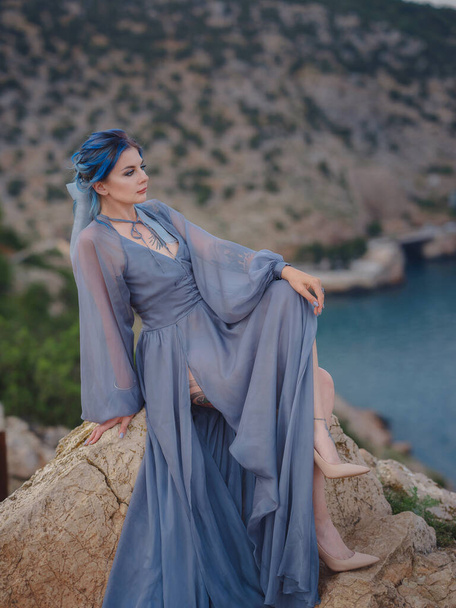 Jonge mooie vrouw met blauw haar in grijze jurk kijkend naar de zee bergen. Vrouwe in de natuur. Modefoto 's. zee nimf wind luisteren wind - Foto, afbeelding