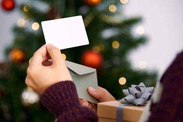 Main tenant maquette carte de voeux de Noël et boîte cadeau pour la conception d'invitation sur fond d'arbre de Noël. - Photo, image