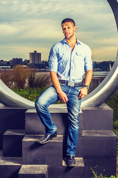 Man Waiting for You. Wearing a light blue shirt, blue jeans, leather shoes, a young handsome guy with a little beard, mustache is standing by a ring structure, looking forward, symbolic for engagement - Foto, Imagen
