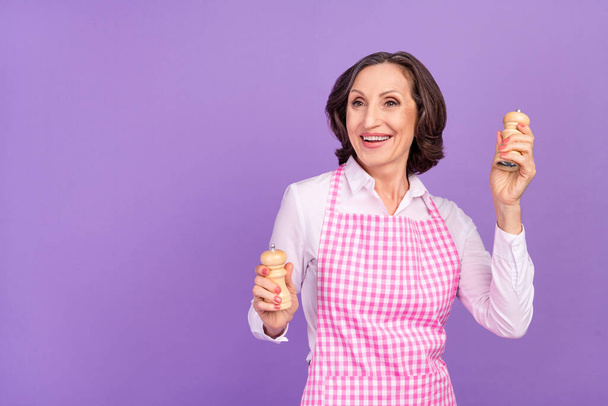 Mutlu, hayalperest yaşlı kadının elinde tuzlu biber tutarken çekilmiş fotoğrafı. Boş görünüyor. Mor arkaplanda izole edilmiş yemek pişiriyor. - Fotoğraf, Görsel