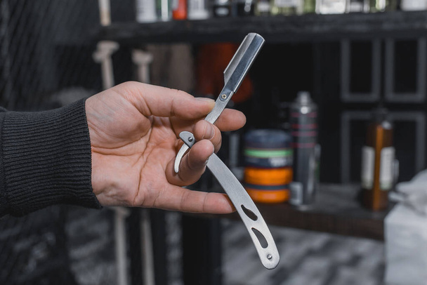 The hand of a barber or professional hairdresser holds a straight tool sharp razor equipment for shaving and beard care, close-up. - Photo, Image