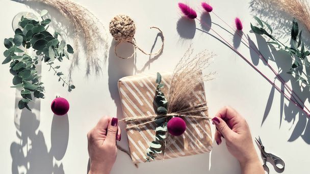 Eco friendly decorations - recycled craft wrapping paper, dry pampas grass, fresh natural eucalyptus twigs and dried bunny tails. Wrapped Xmas gift box with natural Christmas decor in female hands. - Фото, изображение