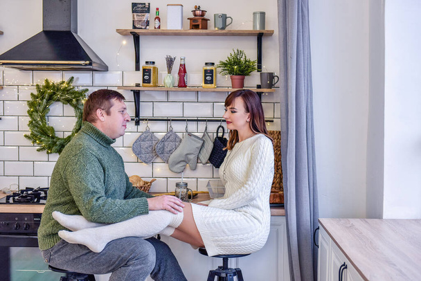 Loving couple in knitted sweaters hugging in fashionable kitchen in stylish interior. Scandinavian style, Christmas decoration. Green spruce wreath hangs over the slab. Festive morning of happy family - Foto, Imagem